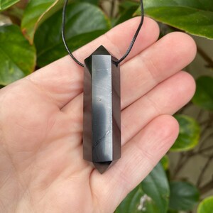 Könnte beinhalten: Schwarzer Obsidian-Doppelspitzen-Anhänger an einer schwarzen Kordelkette. Der Anhänger ist glatt und poliert, mit einem leicht schillernden Glanz.