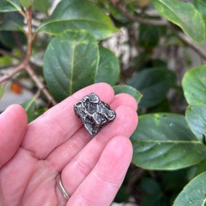 May include: A small, irregular, metallic-coloured object with a textured, pitted surface. The piece has a dark grey, weathered appearance with deep crevices and raised ridges, resembling a fragment of a meteorite or a piece of raw, cast metal.