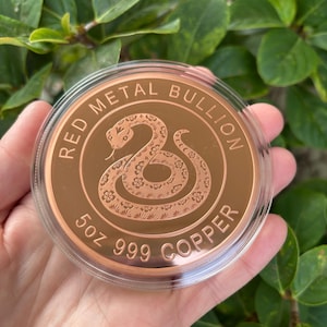 May include: A round copper bullion coin encased in a clear plastic protective capsule. The coin features an embossed snake design in the centre, with the text RED METAL BULLION, 5oz, and 999 COPPER inscribed along the circular border.