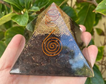 May include: A dark pyramid-shaped orgone energy generator. The pyramid has a clear resin exterior with a dark interior filled with brown and gold flecks. Copper wire spirals are embedded within the resin.