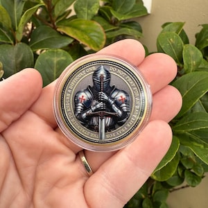 May include: A circular coin depicting a knight in full armour, holding a sword. The knight's armour has red crosses. The coin's edge has text: "THE STRENGTH TO ENDURE A DIFFICULT ONE DO NOT PRAY FOR AN EASY LIFE PRAY".