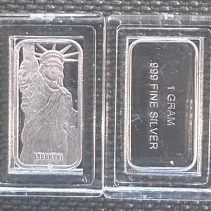 Peut inclure: Deux lingots d'argent dans des étuis en plastique transparent. L'un des lingots représente la Statue de la Liberté avec le mot "LIBERTY" en dessous. L'autre lingot est estampillé "1 GRAMME" et ".999 FINE SILVER".