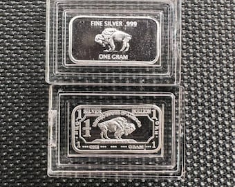 Op de afbeelding: Twee zilveren staven met een buffelontwerp. De bovenste staaf heeft de tekst "FINE SILVER .999 ONE GRAM". De onderste staaf heeft de tekst "1 SILVER UNITED STATES OF AMERICA BULLION 1 ONE GRAM .999".