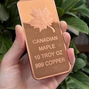 May include: A copper bar with a maple leaf design and the words "CANADIAN MAPLE 10 TROY OZ 999 COPPER" engraved on it. The bar is rectangular and held in a hand, with a green leafy background.