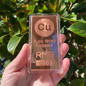 Może przedstawiać: 5-uncjowa sztabka miedzi 999 w przezroczystym plastikowym etui. Sztabka jest miedziana i ma symbol chemiczny miedzi, Cu, oraz liczbę atomową 29. Sztabka ma również napis "RMB 0003".