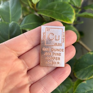 Może przedstawiać: Miedziany sztabka z symbolem pierwiastka Cu, numerem 29 i napisem "ONE OUNCE .999 FINE COPPER USA". Sztabka zawiera również temperaturę topnienia 1085°C i gęstość 8,92 g/cm3.