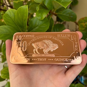 May include: A copper bullion bar inscribed with "10 Copper United States of America Bullion 10" and "10...Ten...Troy...Ounce...10". The bar features an image of a buffalo and the words "In God We Trust".