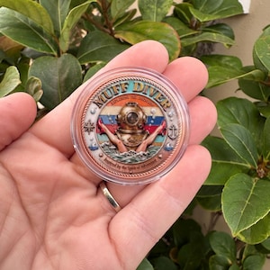 May include: A round, copper-colored coin with the words "MUFF DIVER" and "EXPLORING THE UNKNOWN" around the edge. The coin features a vintage diving helmet, hands, and the text "Inspired by the Spirit of Adventure."