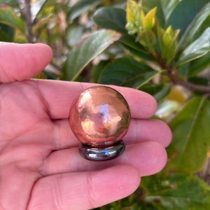 May include: A copper sphere with a hammered texture sits on a dark, metallic ring base. The sphere reflects light, creating highlights. The background features green foliage.