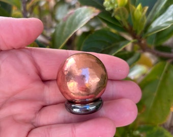 May include: A copper sphere with a hammered texture sits on a dark, metallic ring base. The sphere reflects light, creating highlights. The background features green foliage.