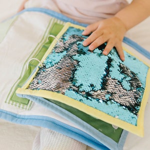May include: A blue and silver sequin book with a yellow border. The book is open and the pages are made of fabric.