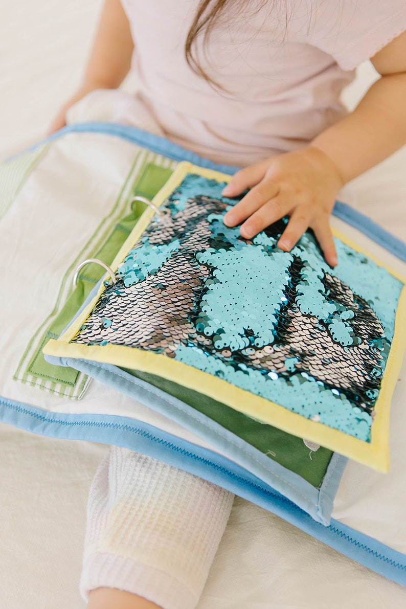May include: A blue and silver sequin book with a yellow border. The book is open and the pages are made of fabric.