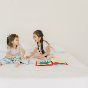 May include: Two young girls are sitting on a white bed, each holding a colorful book. They are smiling and looking at each other. The books are open and show different illustrations.