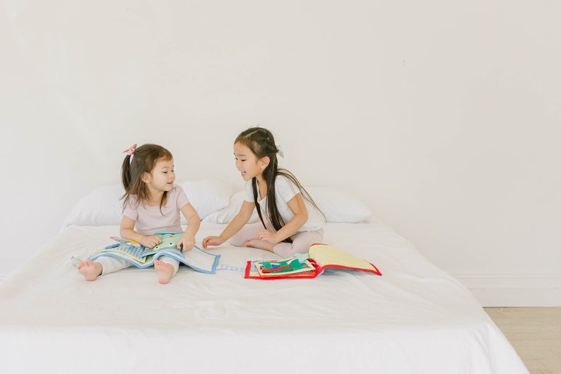 May include: Two young girls are sitting on a white bed, each holding a colorful book. They are smiling and looking at each other. The books are open and show different illustrations.