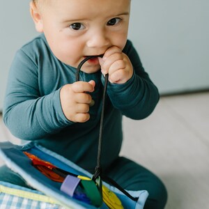 May include: A baby in a blue onesie is playing with a colorful activity toy. The toy has a variety of textures and shapes for the baby to explore.