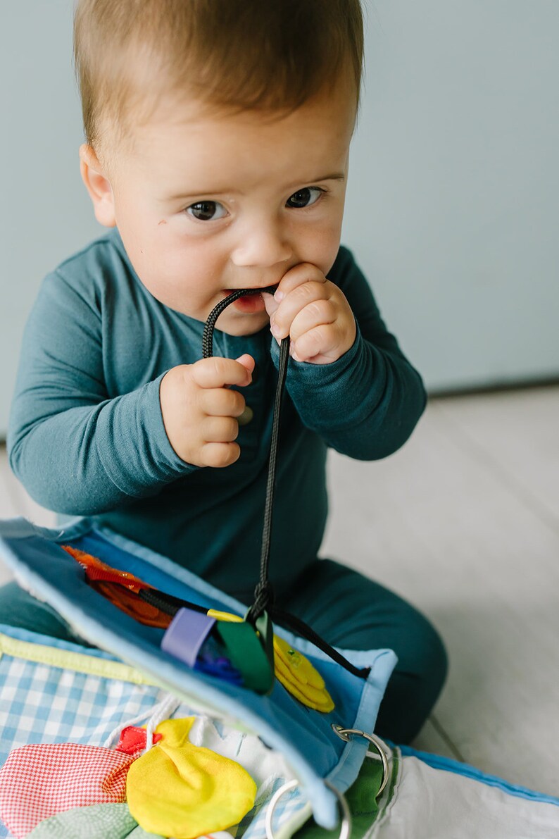 May include: A baby in a blue onesie is playing with a colorful activity toy. The toy has a variety of textures and shapes for the baby to explore.
