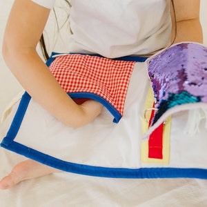 May include: A white fabric book with a red and white checkered pocket, a purple sequin flap, and a red and yellow zipper. The book is being held by a child.