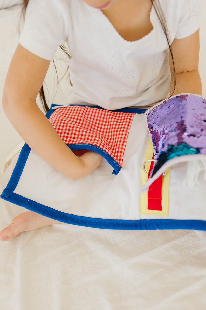 May include: A white fabric book with a red and white checkered pocket, a purple sequin flap, and a red and yellow zipper. The book is being held by a child.