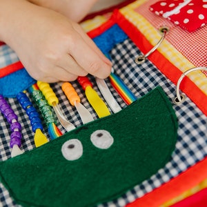May include: A colorful fabric book with various textures and activities for children. It features a green felt monster with eyes, a rainbow ribbon, and colorful beads that can be threaded.
