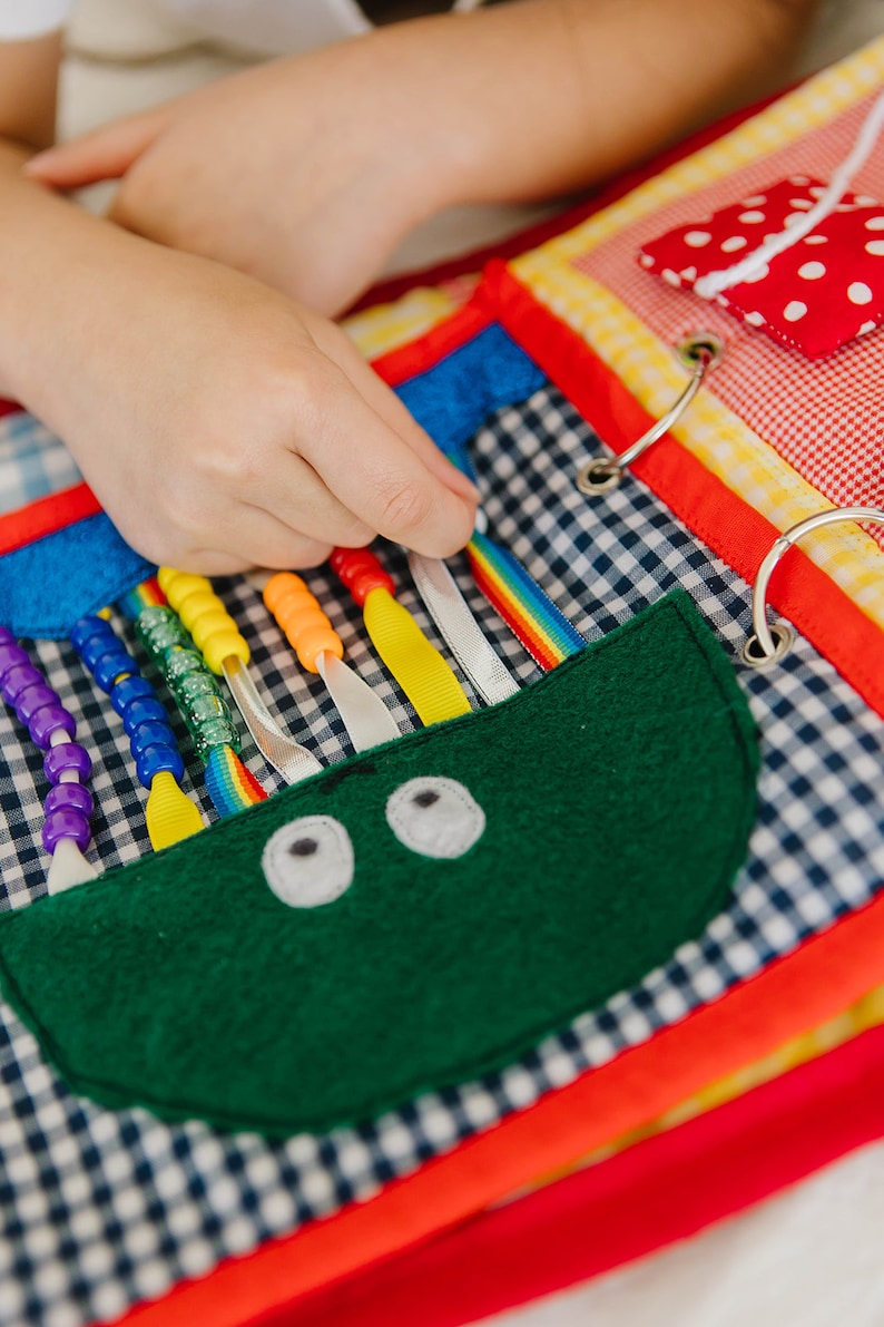 May include: A colorful fabric book with various textures and activities for children. It features a green felt monster with eyes, a rainbow ribbon, and colorful beads that can be threaded.