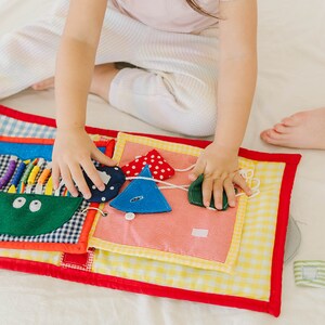May include: A colorful fabric book with various textures and shapes for a child to explore. The book features a green pocket with two eyes, a blue triangle with white polka dots, a blue triangle with a button, and a green leaf shape.