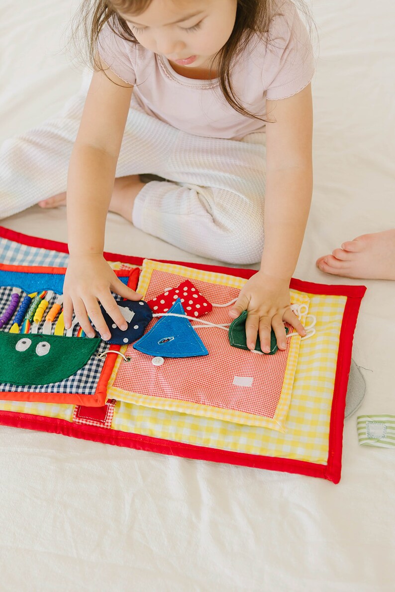 May include: A colorful fabric book with various textures and shapes for a child to explore. The book features a green pocket with two eyes, a blue triangle with white polka dots, a blue triangle with a button, and a green leaf shape.