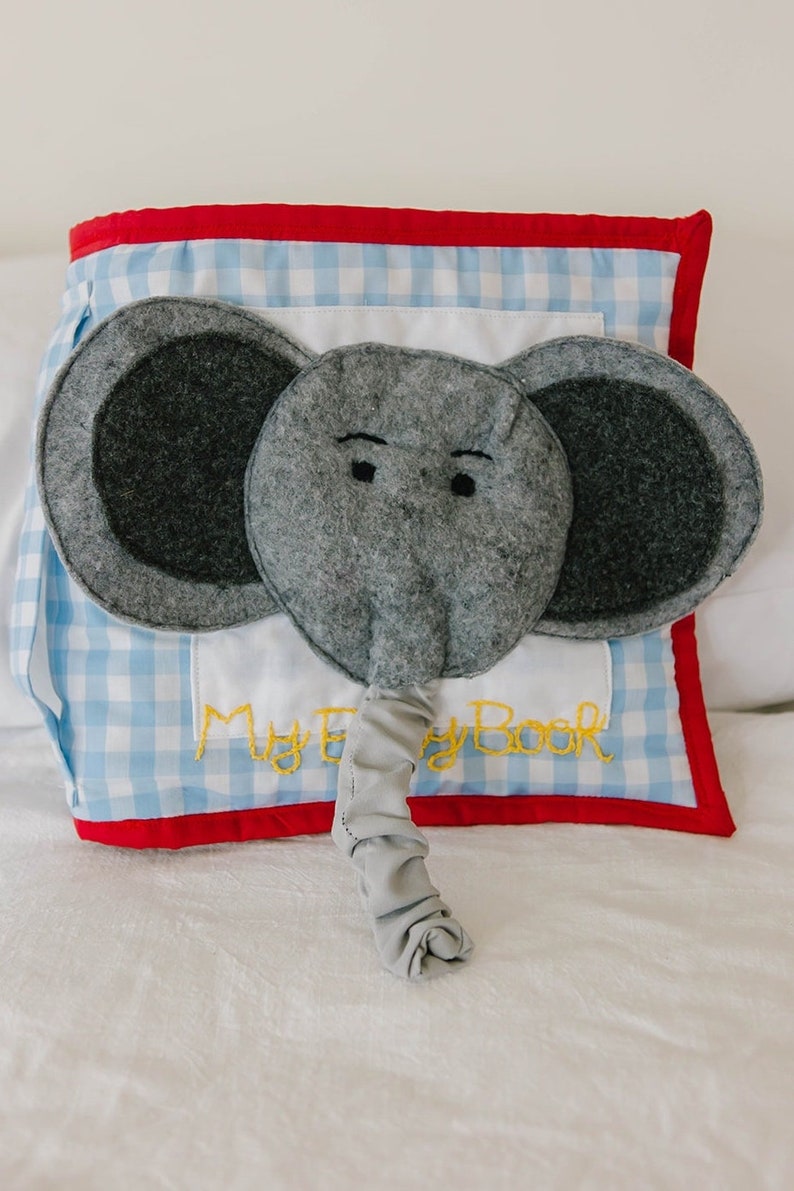 May include: A gray felt elephant with a blue and white gingham book cover. The book cover has the text "My Baby Book" embroidered in yellow thread.