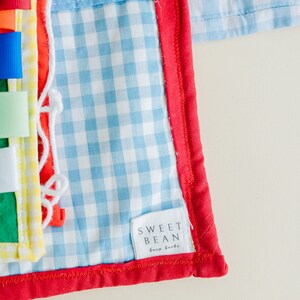 May include: A blue and white gingham fabric baby activity book with a red border and a white tag that says "SWEET BEAN busy books".