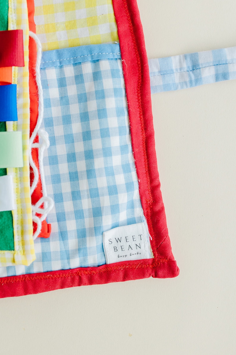 May include: A blue and white gingham fabric baby activity book with a red border and a white tag that says "SWEET BEAN busy books".