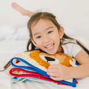 May include: A young girl with long dark hair smiles while holding a stack of colorful fabric books with a lion face on the top book.