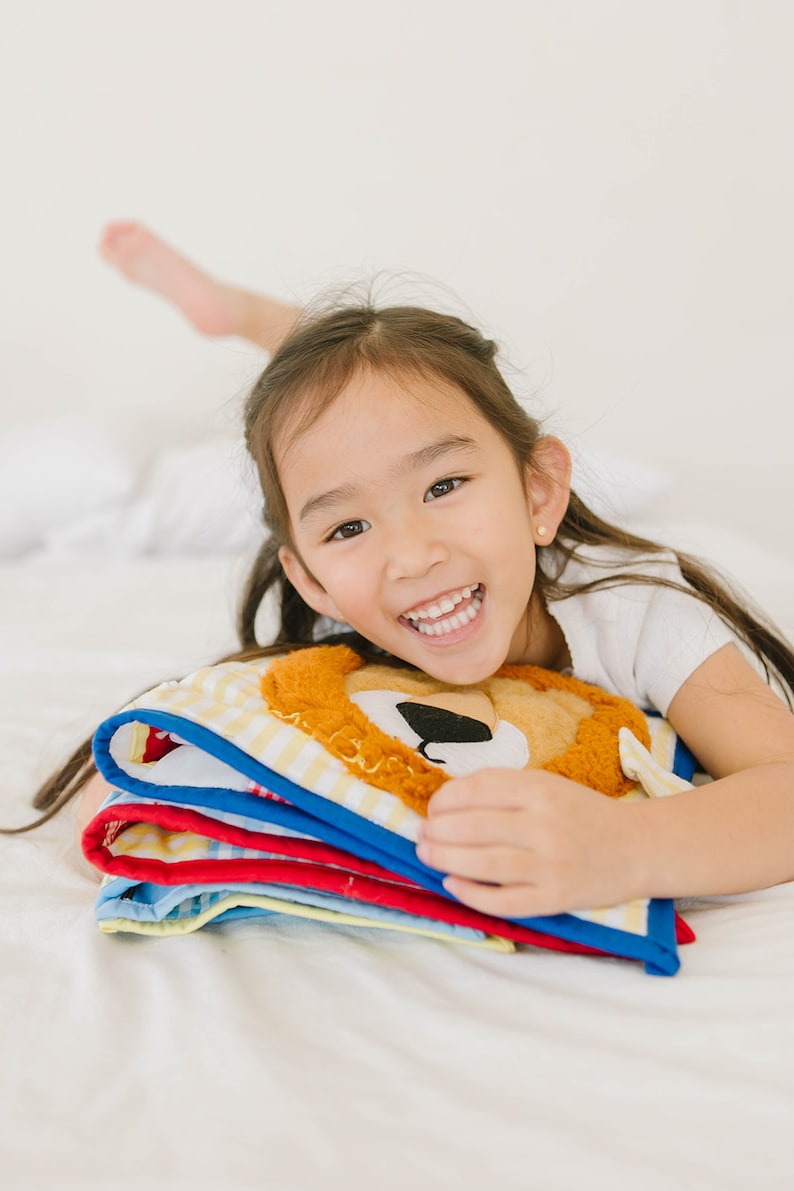 May include: A young girl with long dark hair smiles while holding a stack of colorful fabric books with a lion face on the top book.
