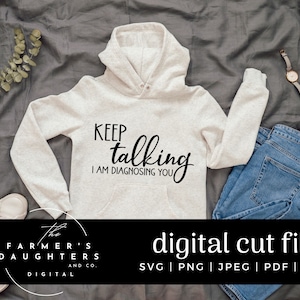 May include: A white hoodie with the text "KEEP talking I AM DIAGNOSING YOU" in black script font. The hoodie is lying on a grey fabric surface with a pair of jeans and a watch.
