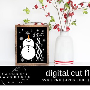 May include: A black sign with white text and a snowman graphic that says "Let it Snow". The sign is on a wooden frame and is sitting on a white surface.