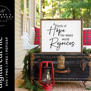 Puede incluir: Un letrero de madera blanco con letras negras que dice "A thrill of Hope the weary world Rejoices". El letrero está apoyado en un baúl de madera negro con herrajes de latón. El baúl está sobre una alfombra tejida frente a un columpio de porche blanco con almohadas de cuadros rojos y negros.