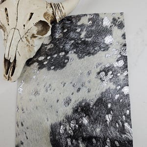 May include: A cowhide piece with a silver metallic pattern over a black and white spotted design. A decorative cow skull with horns is in the background. The cowhide has a soft, textured appearance.