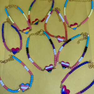 May include: A set of eight beaded bracelets with heart charms featuring different pride flags. The bracelets are made with colorful beads and gold clasps.