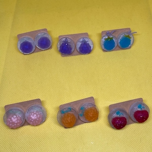 Puede incluir: Seis pares de pendientes de botón con diferentes diseños de frutas. Los pendientes se muestran en pequeñas tarjetas blancas. Los diseños de frutas incluyen uvas moradas, arándanos azules, frambuesas rosadas y melocotones naranjas.