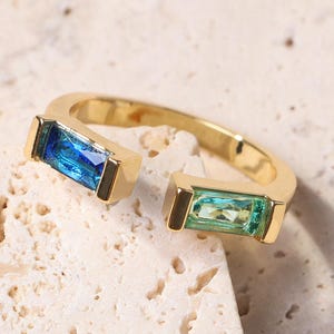 Puede incluir: Un anillo abierto dorado con dos gemas rectangulares. Una es azul vibrante, la otra verde claro. El anillo está sobre un fondo neutro y texturizado.
