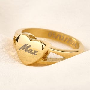 May include: Gold heart-shaped ring with the name "Max" engraved on the front. The ring has a simple band and is a delicate piece of jewellery.