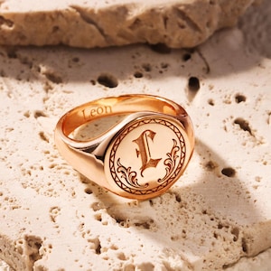 Könnte beinhalten: Roségoldener Siegelring mit dem eingravierten Namen "Leon" auf dem Band. Der Ring zeigt einen eingravierten Buchstaben "L" in einem dekorativen kreisförmigen Rahmen. Der Ring wird auf einer strukturierten, hellfarbigen Oberfläche präsentiert.