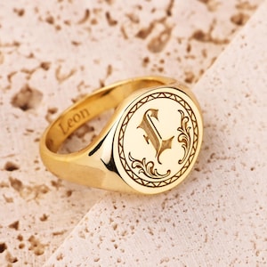 Puede incluir: Un anillo de sello dorado con la inicial "L" grabada dentro de un óvalo decorativo. El anillo tiene el nombre "Leon" grabado en la banda. El anillo está sobre una superficie clara y texturizada.