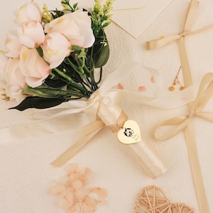 Peut inclure: Un bouquet de roses rose pâle et de verdure, noué avec un ruban crème. Un charm en forme de cœur doré avec une illustration noire et du texte est attaché. D'autres rubans et un petit cœur tressé sont également visibles.
