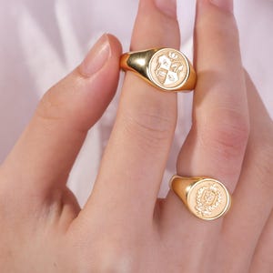 May include: Two gold signet rings with detailed crest designs. One ring is on a finger, the other is held by a hand. The rings have a polished finish and a classic, elegant style. The crests feature intricate patterns.