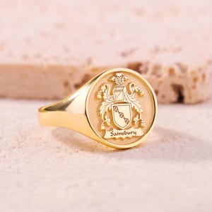 Puede incluir: Un anillo de sello dorado con un diseño de escudo detallado. El anillo presenta una cara redonda con un escudo familiar y el nombre "Sainsbury" grabado debajo. El anillo está sobre un fondo neutro.