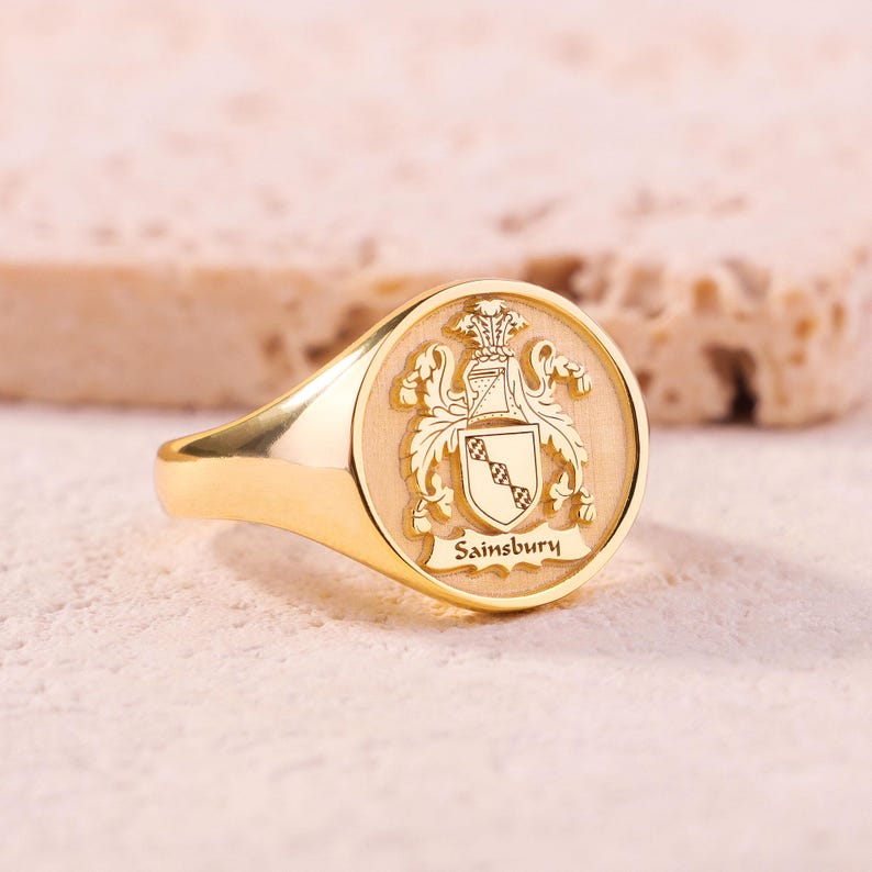 May include: A gold signet ring featuring a circular face with an engraved heraldic crest. The design includes a helmet, ornate scrollwork, a shield with a diagonal checkered band, and the name Sainsbury inscribed on a banner at the bottom.