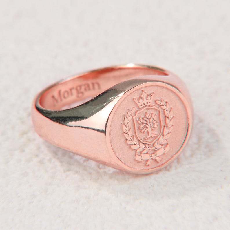 May include: A rose gold signet ring featuring a circular face engraved with a crest depicting a tree, a crown, and laurel branches. The side of the ring band is engraved with the name Morgan in a clear, sans-serif font, showcasing a polished finish.