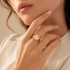 May include: A gold signet ring featuring a circular face with an engraved heraldic crest design. The ring has a polished metallic finish and a smooth, tapered band that sits comfortably on the finger, showcasing intricate detailing on the face.