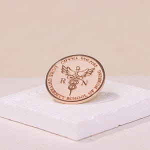 以下が含まれることがあります： 縁に「DOVE UNIVERSITY SCHOOL OF NURSING」と「CHYNNA BSN 2018」の文字が刻まれた金色のピン。ピンの中央には、医学の象徴であるカドゥケウスが描かれており、両側に「RN」の文字が刻まれています。