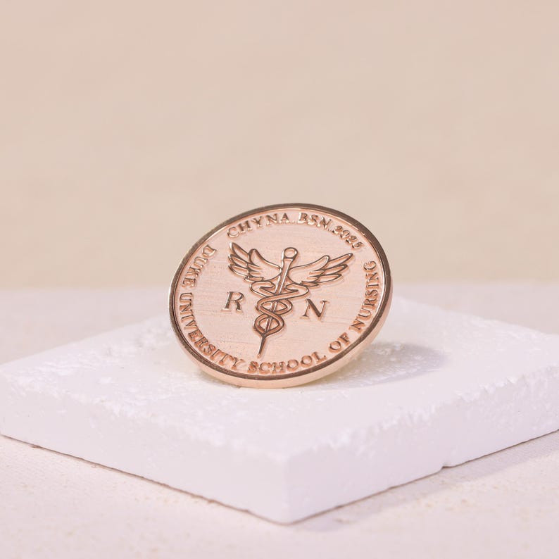 以下が含まれることがあります： 縁に「DOVE UNIVERSITY SCHOOL OF NURSING」と「CHYNNA BSN 2018」の文字が刻まれた金色のピン。ピンの中央には、医学の象徴であるカドゥケウスが描かれており、両側に「RN」の文字が刻まれています。
