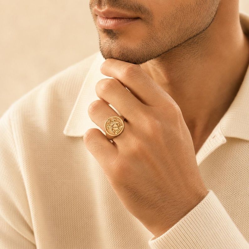 May include: A gold signet ring featuring a circular face with an engraved crest design. The ring has a polished metallic finish and a smooth band, worn on the ring finger. The design on the face includes intricate detailing and a central emblem.